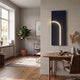 A cozy, modern home office features a wooden desk, leather chair, and a potted plant by a large window. Sunlight highlights Giant Sculptures' Luminarch Navy Curve LED Minimalist Wall Art on the light gray wall with contemporary lighting.