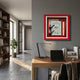 A modern home office features a wooden desk, black chair, lamp, and books. Vintage flair comes from the Cavendro Newspaper & Pipe Framed Wall Art by Giant Sculptures on a gray wall. Sunlight fills the space with shelves and a potted plant nearby.