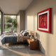 A modern bedroom with a neatly made bed, a wooden nightstand with a lamp, large sunlit windows, and Cavendro Single Cigar & Torch Lighter Framed Wall Art by Giant Sculptures in a striking red frame on the wall.