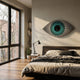 A modern bedroom with large windows, wooden floors, and a low bed features the Giant Sculptures Aurevion Teal Radiant Eye 3D Wall Art above the headboard. Sunlight highlights bookshelves and a desk lamp in the airy space.