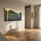 A minimalist room with light wooden floors, beige walls, and sunlight through sheer curtains is accented by the Seralyon Green Drip Peak Wide Canvas Wall Art by Giant Sculptures above a built-in bookshelf filled with books.