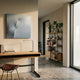 A modern, minimal home office with a wooden desk, black chair, desk lamp, abstract art, and Giant Sculptures’ Veylora Ice Blue Folded Texture Fabric 3D Wall Art on the wall, plus bookshelves with books and plants and large windows.