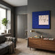 A modern living room with gray walls, a large window, gray sofa, wood floors, sideboard with books and lamp, potted plant, and Giant Sculptures' Loftera Blue Sculpted Fabric & Abstract Canvas 3D Wall Art on the wall.