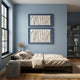 A cozy bedroom with light blue walls, a large window, a neatly made bed with beige bedding, and Giant Sculptures' Foldara Cream & Blue Draped Paper Dual-Panel 3D Wall Art above the bed; shelves with books and decor sit in the corner.