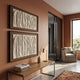 A modern living space with terracotta walls, wabi-sabi decor, two Foldara Cream & Mocha Draped Paper Dual-Panel 3D Wall Art pieces by Giant Sculptures, a wooden console with books and lamp, beige armchair by large windows, and a patterned rug on concrete.