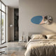 Minimalist bedroom with neutral tones, a low bed with beige bedding, and Giant Sculptures’ Aurelune Blue & Pink Silver Metal Disc 3D Wall Art above the headboard. A floor lamp by large windows illuminates a side table holding books and a vase.