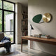 A modern home office with a wooden desk, books, a black lamp, and the Aurelune Green & White Gold Metal Disc 3D Wall Art by Giant Sculptures. Large windows provide natural light, and a gray armchair with a blanket sits near built-in book shelves.