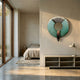 A modern, minimalist bedroom features large windows, a bed with neutral bedding, and a bedside table. Above a low shelf, the Vortaryn Verdigris & Steel Circle 3D Wall Art by Giant Sculptures accents the wall as sunlight streams in for a serene vibe.