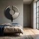 A modern bedroom with a low bed, beige and blue bedding, wooden floors, a patterned rug, sunlight streaming through tall windows, and the Vortaryn Charcoal & Concrete Circle Sculptural 3D Wall Art by Giant Sculptures above the bed.