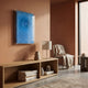 A modern living room with peach walls features a wooden console topped with books and decor, a lamp, a chair with a beige blanket, and Giant Sculptures’ Lumora Deep Blue Acrylic Framed 3D Paper Wall Art above. Sunlight fills the space through large windows.