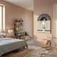 A cozy, modern kids’ bedroom features pink walls, star-patterned bedding, a bookshelf with toys and books, a small wooden rocking chair, and adorable Cuddiloft Cream Bunny Plush Display 3D Wall Art by Giant Sculptures. Sunlight streams in through a large window.