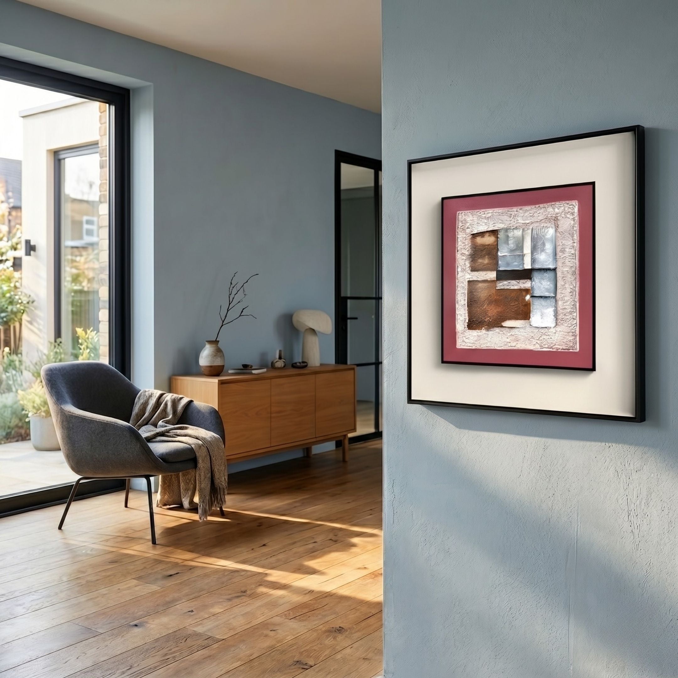 A modern living room with wooden floors, a gray armchair, and a wooden sideboard features minimalist decor. Terralume Rose Clay & Silver Block Textured Framed Wall Art by Giant Sculptures hangs on a pale blue wall near large windows.