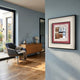 A modern living room with wooden floors, a gray armchair, and a wooden sideboard features minimalist decor. Terralume Rose Clay & Silver Block Textured Framed Wall Art by Giant Sculptures hangs on a pale blue wall near large windows.