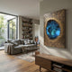 Vorthera Sapphire Burst Textured Gold 3D Wall Art with a jagged blue crystalline center, displayed in a modern living room with a grey sofa and bookshelf.