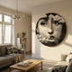Lunoria Cloud Face Round Wood Embossed Wall Art displayed on a cream living room wall above a grey sofa, wooden coffee table, and bookshelf.