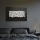 Stravira Black I Sculptural White Metal 3D Wall Art with interwoven white iron strips on a black panel, mounted above a dark grey bed and nightstand.