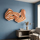 Riborra Orange Layered Wood Carving 3D Wall Art by Giant Sculptures, mounted on a blue wall above a console with books, vase, and lamp beside a grey armchair.