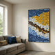 Tilora II Abstract Folded Leather 3D Wall Art by Giant Sculptures in blue, yellow, white and brown, framed in black above a grey sofa with cushions.