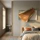 Valmeris Amber Fold Sculptural Metal 3D Wall Art mounted above a wooden bed with linen pillows in a modern bedroom with pale grey walls and oak flooring.