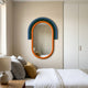 A modern bedroom features a neatly made bed with white bedding, gray and mustard pillows, a bedside table with a lamp, and the Arcaluna Blue & Orange Layered Arch Mirror 3D Wall Art by Giant Sculptures on a beige wall.