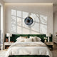 A modern bedroom features a green velvet headboard, white bedding, two bedside tables with lamps and flowers, and the Iriselle Amber Flare Crystal Porcelain Round 3D Wall Art by Giant Sculptures above the bed. Sunlight casts window-frame shadows on the wall.