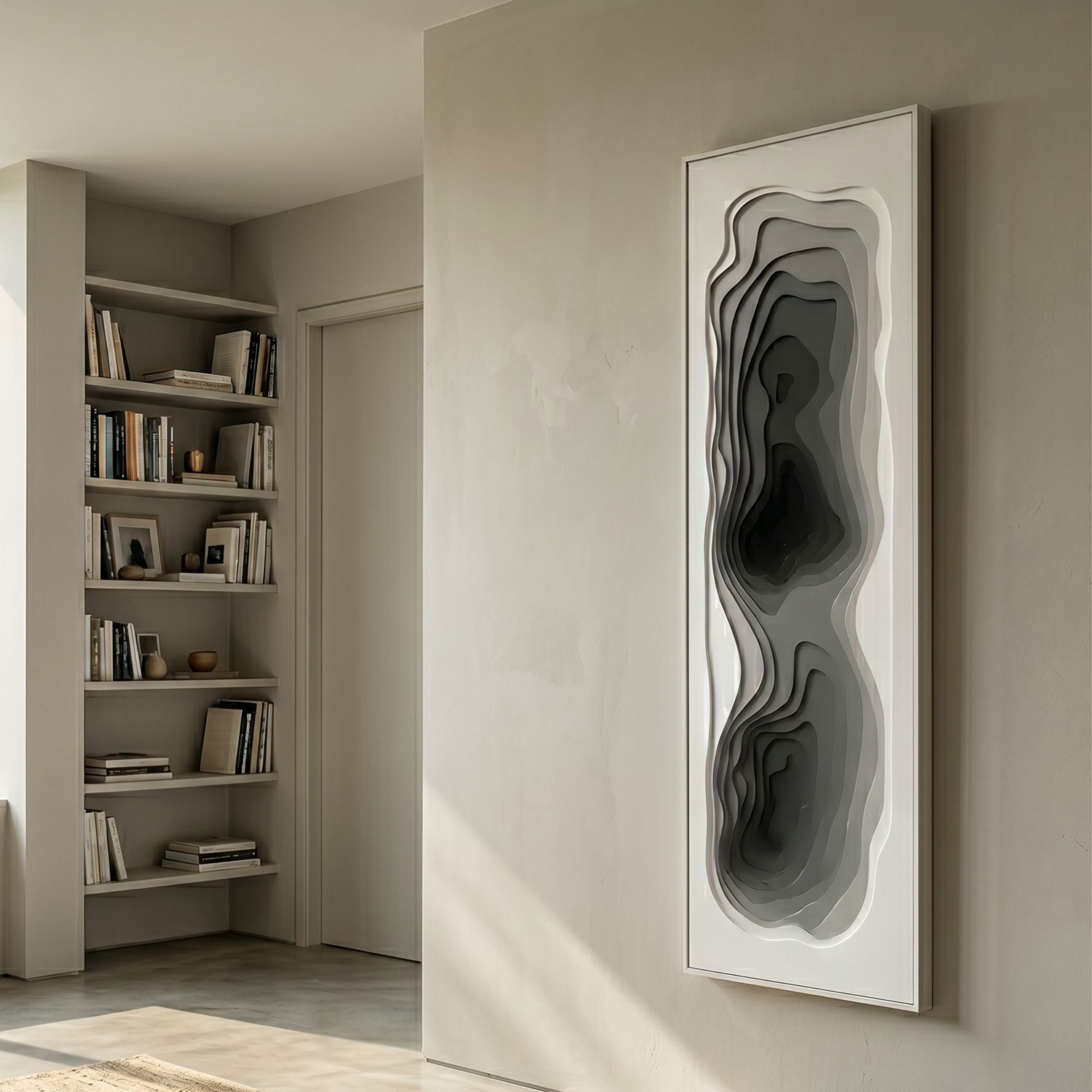 Bathys Grey Gradient Carved 3D Wall Art mounted on a pale wall beside a built-in bookshelf, featuring layered wavy carved forms in graduating grey tones within a tall white frame.