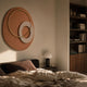 Orbien Terracotta & Gold Ring Sandstone Mural Wall Art mounted above a bed in a warm-toned bedroom with a mushroom lamp and dark wood bookshelf nearby.