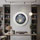 A modern office features a marble desk, chair, plants, and sculptures. The highlight is the Iriselle Frost Grey Crystal Porcelain Round 3D Wall Art by Giant Sculptures, resembling an eye on a gray paneled wall, with warm lighting enhancing the elegant decor.