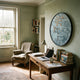 A cozy home office with a wooden desk, laptop, books, lamp, and coffee mug. By the window is an armchair and modern decor, including the Giant Sculptures Florivonne Gilded Metal Leaf Round 3D Wall Art on a pale green wall.