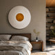 Ventus Amber Glow Wood Carving 3D LED Wall Art mounted above a wooden bed, its white rippled disc and glowing amber centre lighting the neutral bedroom.