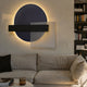Aeon Onyx Graphite Leather 3D LED Wall Art by Giant Sculptures with circle, black bar, and lined square, backlit above a beige sofa beside a bookshelf.