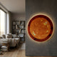 Galaxia Sun Crystal Porcelain LED Wall Art glowing amber and red on a grey wall beside a modern living room with a beige sofa and bookshelf.