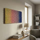 Spectrum Weave Horizontal Sunset Hues Wood Relief 3D Wall Art by Giant Sculptures, gradient yellow to pink, red and blue, mounted above a console with lamp and books.