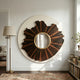 The Lunestre Bronze Ripple Organic Round Mirror 3D Wall Art by Giant Sculptures hangs on a white wall in a bright living room, near a brass floor lamp, bookshelves, and a plant. A doorway leads to a cozy sofa in the next room.