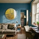 A cozy living room with blue walls, a beige sofa, wooden desk and chair by a large window, potted plants, books, a lamp, and the Auralune Golden Sketch Abstract Round 3D Wall Art by Giant Sculptures adding a striking touch.