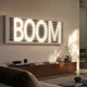 Aurova I Contemporary Glow 3D LED Wall Art by Giant Sculptures displaying illuminated white BOOM lettering behind grey horizontal slats, mounted above a walnut sideboard.