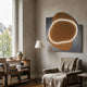 Gleam Russet Harmony 3D Leather LED Wall Art with a russet leather form and white LED curve over grey panels, displayed above an armchair in a living room.