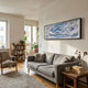 Fluenta Blue Ripple Linear Cardboard 3D Wall Art by Giant Sculptures in a black frame above a grey sofa in a sunlit living room with bookshelf.
