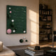Orbison Green 3D Wood Relief Wall Art with floating white, pink, and green spheres displayed in a sunlit living room beside a dark bookshelf.