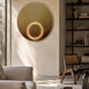 Aurora Golden Halo Crystal Porcelain LED Wall Art mounted on a cream wall in a modern living room with a cane chair, dark bookshelf, and beige sofa.
