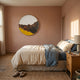 A cozy bedroom with pink walls, a neatly made bed in beige and blue, a wooden nightstand with books, slippers on a woven rug, and the Auralune Golden Ridge Abstract Round 3D Wall Art by Giant Sculptures above the bed. Sunlight filters through sheer curtains.