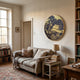 A cozy living room features a beige sofa with a patterned pillow and throw blanket. Above, the Auralune Golden Market Scene Round 3D Wall Art by Giant Sculptures adds flair. A side table with books and a cup plus bookshelves and a large window complete the space.