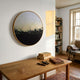 The Auralune Dusk Line Gradient Abstract Round 3D Wall Art by Giant Sculptures hangs on a white wall above a wooden desk with books and pens. Sunlight streams in, illuminating the cozy corner with a chair and bookshelf.