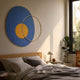 Solaro Radiant Blue & Yellow Round Mural Wall Art by Giant Sculptures displayed above a wooden bed with grey linens, beside a mushroom lamp and rubber plant.