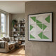 Prismatics Green Geometric Cardboard Wall Art by Giant Sculptures in a black frame, displayed on a beige wall in a modern living room with a linen sofa and bookshelf.