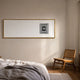 Lustreline Black & White Right Plaster Statue Cardboard Wall Art in a gold frame mounted above a bed beside a cane chair in a minimalist bedroom.