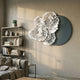 Fleur Halo Grey Round White Metal Floral Decorative Wall Art mounted in a modern living room above a bookshelf and grey sofa, with layered white petals on a teal-grey disc.
