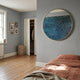 A cozy bedroom with light blue walls, a round mirror, wooden floor, rust bedding, framed photos, and the Auralune Blue Horizon Abstract Round 3D Wall Art by Giant Sculptures above the bed; plus a closet door and woven laundry basket by a sunlit window.