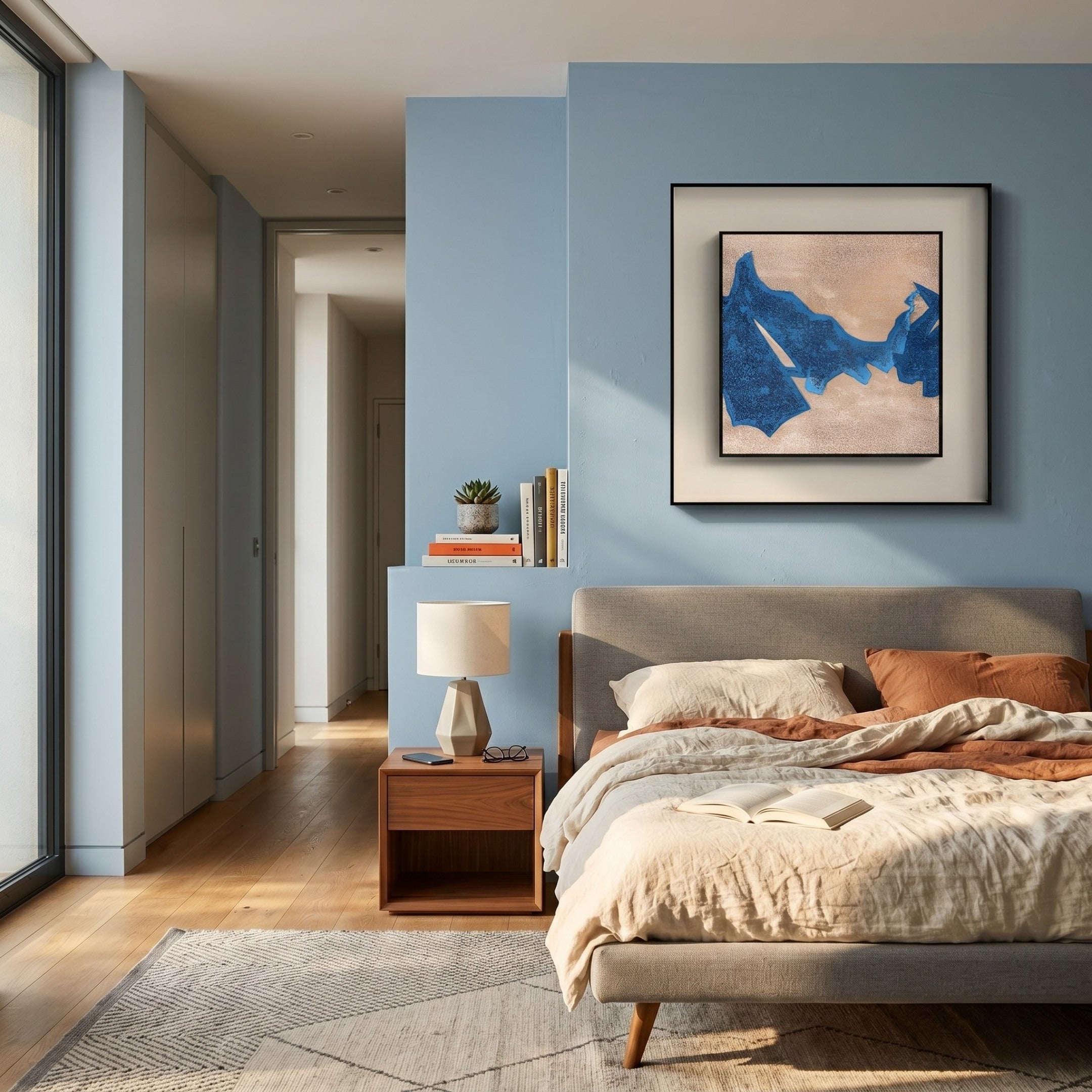 A cozy bedroom with light blue walls, a large bed with beige linens, a wooden nightstand, and a small potted plant. Sunlight highlights the Stratum Blue Crest Textured Abstract Framed Wall Art by Giant Sculptures above the bed.