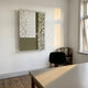 A minimalist room with wooden floors, a table, and a black chair by a small bookshelf under a window features Giant Sculptures’ Texturien White & Olive Paper Abstract 3D Wall Art on the wall, illuminated by soft sunlight.